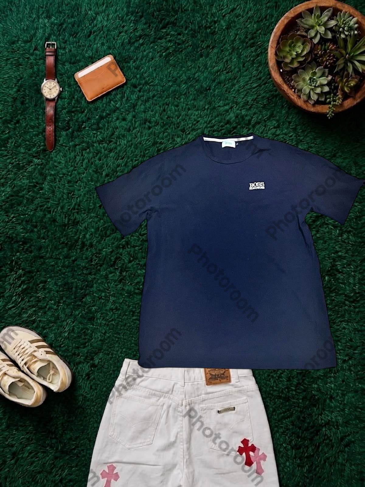 Result from a photo SaaS: navy blue T-shirt on artificial turf, with a watch, a wallet, a plant, sneakers and a pair of trousers around it, and its watermark on top.